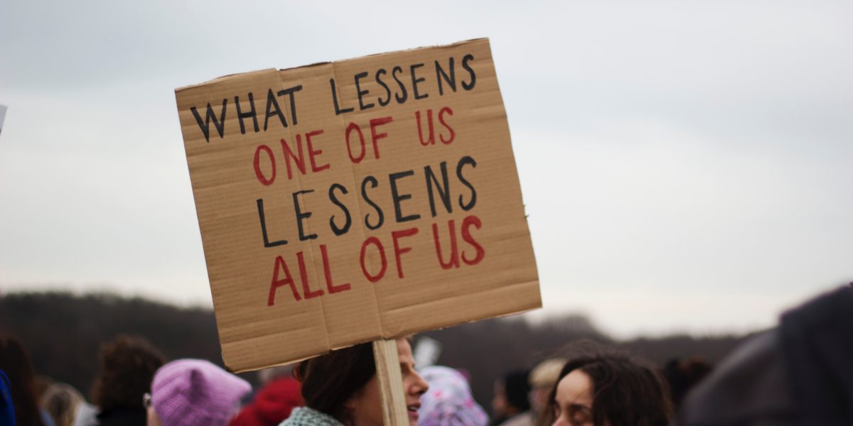 Una donna durante una manifestazione regge un cartello con la scritta "What lessens one of us lessens all of us".