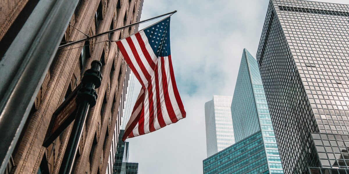 Immagine di una bandiera statunitense in primo piano circondata da grattacieli. 
